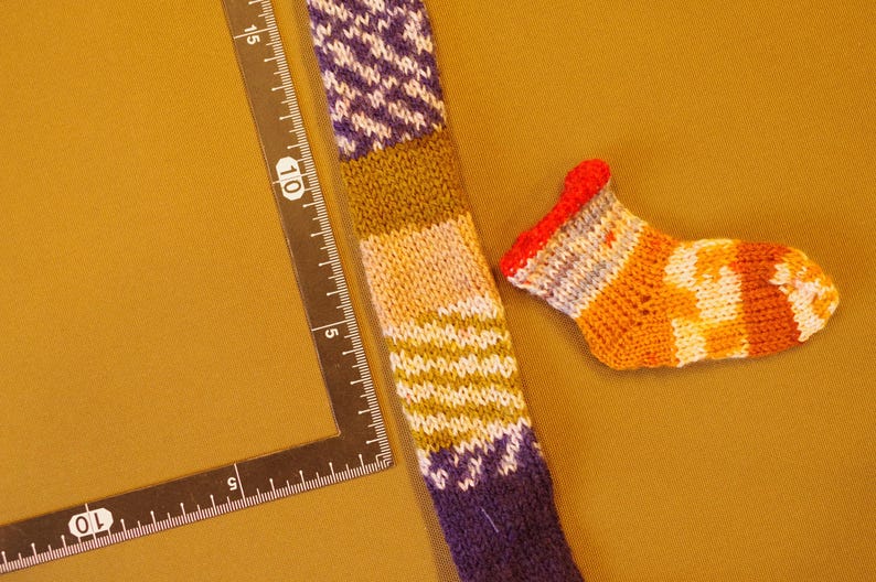 K&ouml;nnte beinhalten: Eine bunte gestrickte Socke mit einem lila, gr&uuml;nen und wei&szlig;en Muster. Eine kleinere, gestreifte Socke mit orange, wei&szlig;en und roten Streifen ist ebenfalls abgebildet.
