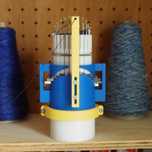 K&ouml;nnte beinhalten: Eine blaue und gelbe Strickmaschine mit mehreren Nadeln. Die Maschine steht auf einer Holzoberfl&auml;che.