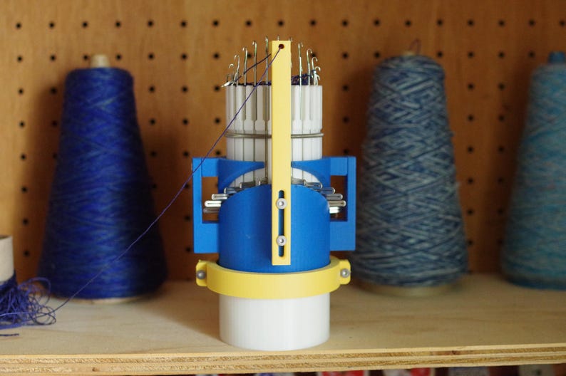 K&ouml;nnte beinhalten: Eine blaue und gelbe Strickmaschine mit mehreren Nadeln. Die Maschine steht auf einer Holzoberfl&auml;che.