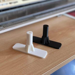 May include: Two plastic knitting machine parts. One is white, the other is black. Both have a rectangular base and a cylindrical post. They are sitting on a light brown wooden surface.
