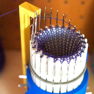 Può includere: Primo piano di un tessuto all'uncinetto viola che viene realizzato su un telaio circolare per la maglia con una base gialla e aghi bianchi.