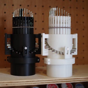 K&ouml;nnte beinhalten: Zwei 3D-gedruckte Nadelhalter, einer schwarz und einer wei&szlig;, mit einem runden Sockel und einer drehbaren Oberseite. Die Halter sind mit Nadeln gef&uuml;llt.