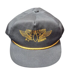 May include: A black baseball cap with a flat brim and a yellow rope detail. The cap features an embroidered design with the words "LONG RANGE BOMBER" and a stylized image of a bomb with wings in yellow.