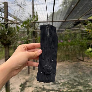 Może przedstawiać: Ciemnoszary, prostokątny obiekt o fakturze drewna z kwiatowym wzorem i małymi okrągłymi akcentami. Obiekt zawieszony jest na cienkim drucie, co sugeruje, że może być elementem dekoracyjnym lub dziełem sztuki. Tło to rozmyty widok na zewnątrz.