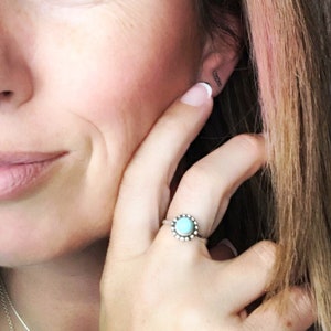 May include: A silver ring with a turquoise stone set in a circle of small beads. The ring is on a person's finger.