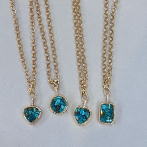May include: Four gold-toned necklaces with different pendant shapes: a triangle, a circle, a heart, and a rectangle. Each pendant is filled with turquoise-colored stone chips. The necklaces hang on a white surface.