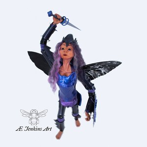 May include: A purple-haired fairy doll with black wings, wearing blue armour and holding a blue dagger. The doll is posed in a fighting stance with one leg raised. The doll is made by AE Jenkins Art.