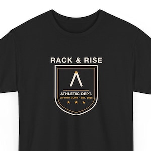 Può includere: Una t-shirt nera a maniche corte con una stampa grafica sul petto. Il design include il testo RACK & RISE sopra un emblema a scudo contenente il testo ATHLETIC DEPT. LIFTING CLUB EST. 2025, un simbolo a chevron e tre stelle.