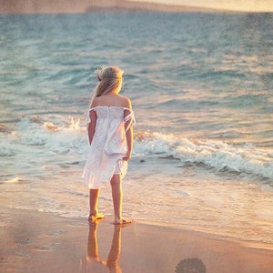May include: A young girl in a white dress stands on a sandy beach, looking out at the ocean. The water is a light blue and the sky is a soft orange. The girl's hair is long and blonde, and she is wearing a flower crown. The image has a vintage, faded look.