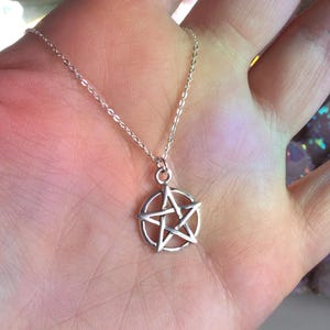 May include: A silver-colored necklace featuring a pentagram pendant. The pendant is a five-pointed star within a circle. The necklace is displayed on a hand, with a soft, colorful background.