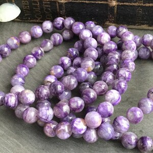 May include: Several purple charoite gemstone bracelets. The bracelets are made of round, polished beads with a marbled appearance. The background includes a vintage book and decorative mushrooms, creating a natural, earthy aesthetic.