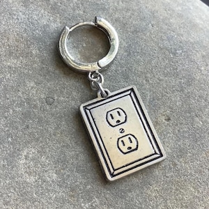 May include: Silver-toned earring featuring a rectangular charm with a detailed electrical outlet design. The charm is suspended from a small hoop earring. The earring is made of metal and has a vintage or industrial aesthetic.