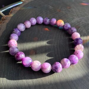 May include: A bracelet made of round, polished stones in shades of purple, lavender, and pink. The stones are strung together, forming a circular shape. The stones have a marbled appearance, with variations in colour and texture.