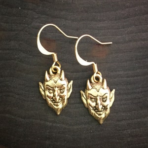 May include: A pair of gold-tone earrings featuring a detailed devil face design. The earrings are made of metal and have a hook closure.