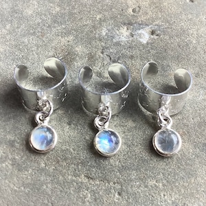 May include: Three silver ear cuffs with a dangling, round, iridescent gemstone. The ear cuffs have a simple, open design and are set against a textured, gray surface. The gemstones have a blue and white color.