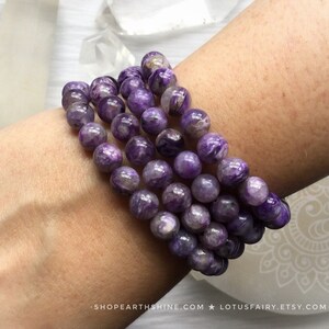May include: A close-up of several bracelets made of round, purple beads. The beads have a marbled appearance with lighter purple and white swirls. The bracelets are stacked on a person's wrist. The background includes crystals and the text "SHOPEARTHSHINE.COM * LOTUSFAIRY.ETSY.COM".
