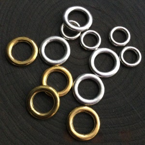 May include: A collection of metal rings in various sizes and finishes. Some rings are gold-toned, while others are silver-toned. The rings are scattered on a dark, textured surface, creating a contrast between the shiny metal and the dark background.