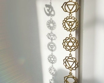 May include: A gold-colored metal hanging mobile with seven charms. The charms are in the shape of the seven chakras, including the third eye, root, sacral, solar plexus, heart, throat, and crown chakras. The mobile also features an Om symbol at the top.