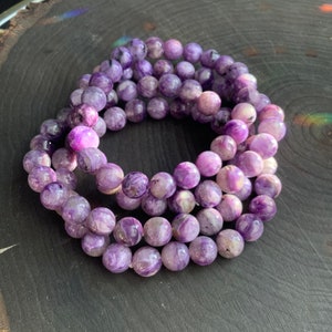 May include: A collection of purple beaded bracelets. The beads are round and have a marbled appearance with shades of lavender, violet, and white. The bracelets are arranged in a circular pattern on a dark wooden surface.