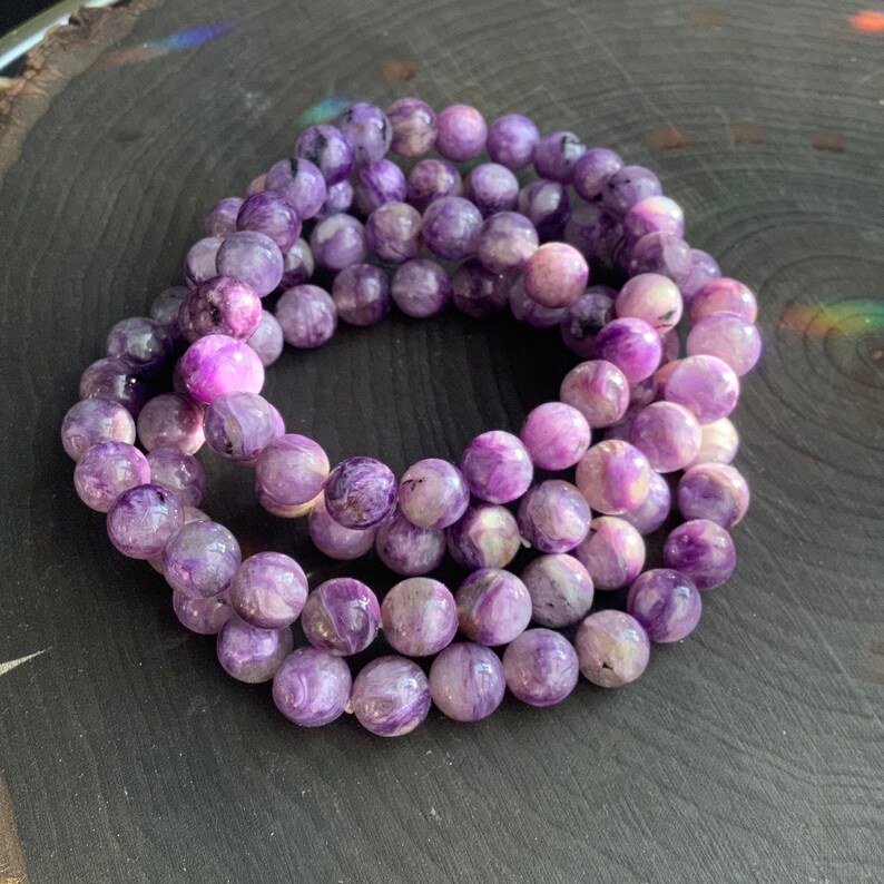 May include: A collection of purple beaded bracelets. The beads are round and have a marbled appearance with shades of lavender, violet, and white. The bracelets are arranged in a circular pattern on a dark wooden surface.