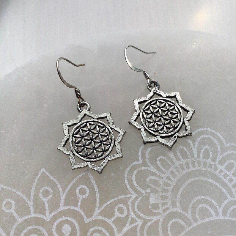 May include: A pair of silver-toned earrings. Each earring features a flower-like design with a geometric pattern at the center. The earrings have a hook closure. The earrings are displayed on a white surface.