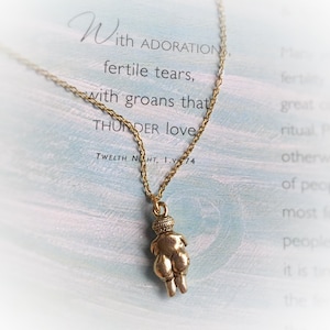 May include: A gold chain necklace with a pendant of a small, stylized female figure. The pendant is made of a gold-toned metal and has a textured surface. The necklace is hanging on a card with the text "With ADORATION, fertile tears, with groans that THUNDER love. TWELTH NIGHT, 1.V.74"