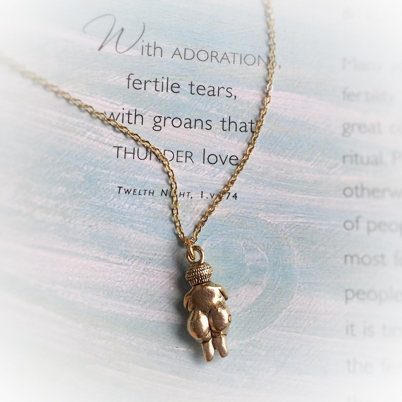 May include: A gold chain necklace with a pendant of a small, stylized female figure. The pendant is made of a gold-toned metal and has a textured surface. The necklace is hanging on a card with the text "With ADORATION, fertile tears, with groans that THUNDER love. TWELTH NIGHT, 1.V.74"