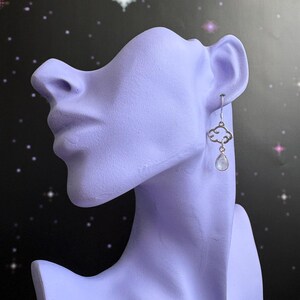 May include: A silver earring with a cloud-shaped design and a small, teardrop-shaped, iridescent stone. The earring is displayed on a lavender-colored bust against a dark background with white and pink stars.