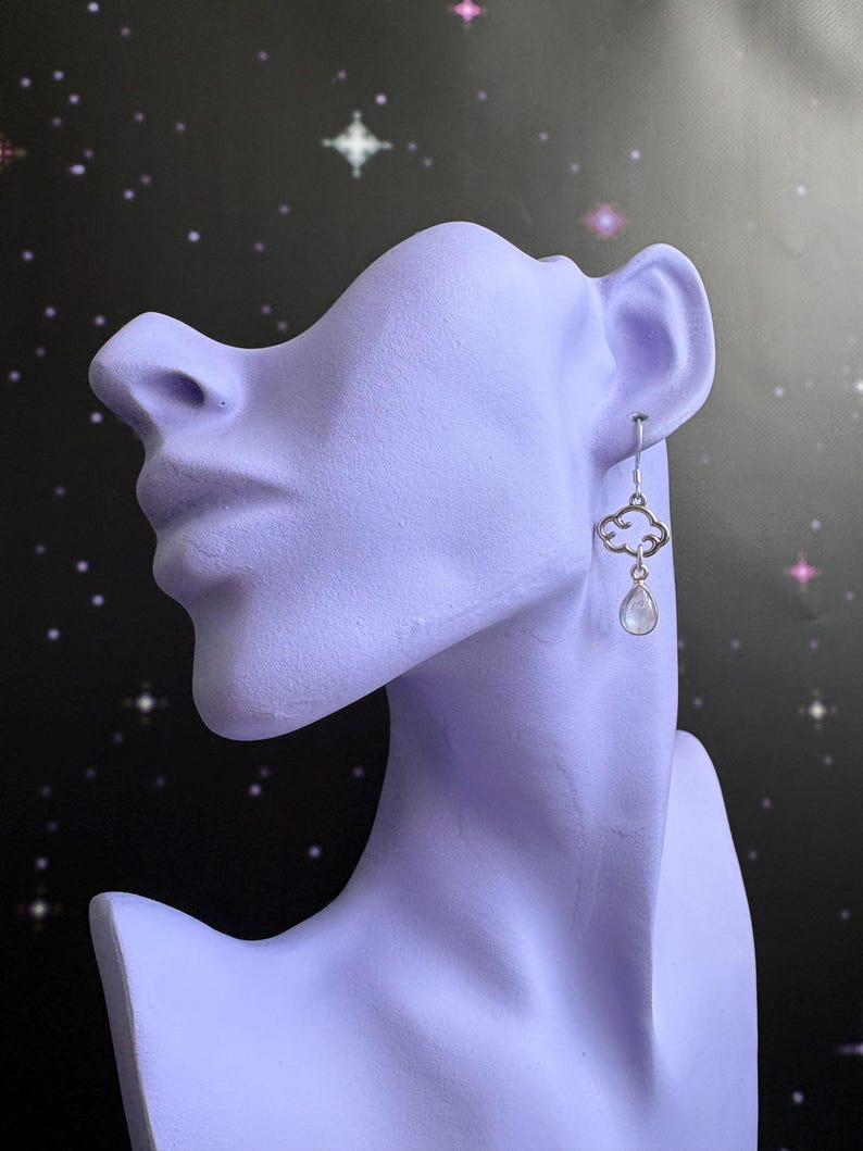 May include: A silver earring with a cloud-shaped design and a small, teardrop-shaped, iridescent stone. The earring is displayed on a lavender-colored bust against a dark background with white and pink stars.