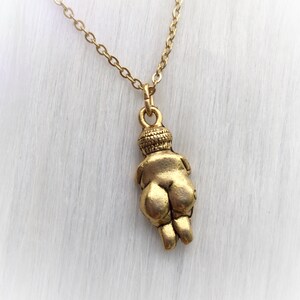 May include: Gold necklace with a pendant of a stylized female figure. The figure is depicted in a seated position with her arms crossed over her chest.