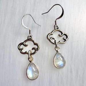 May include: A pair of silver-toned cloud-shaped earrings with teardrop-shaped, iridescent gemstones. The earrings have a hook closure and are displayed against a white, textured background. The gemstones reflect a range of colors.