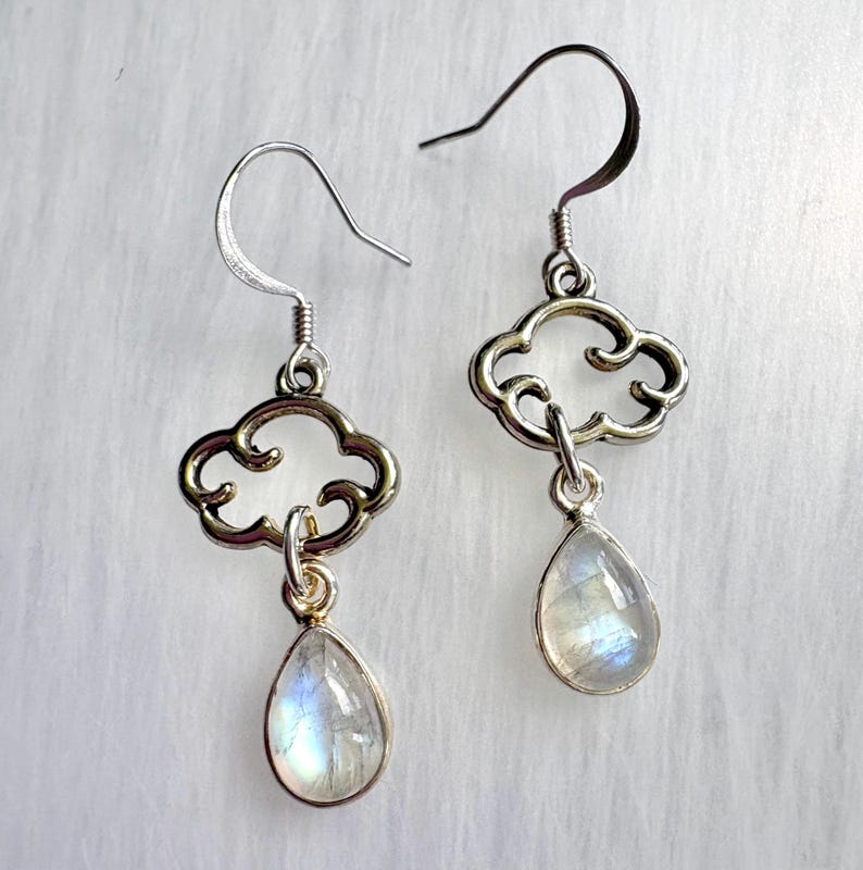May include: A pair of silver-toned cloud-shaped earrings with teardrop-shaped, iridescent gemstones. The earrings have a hook closure and are displayed against a white, textured background. The gemstones reflect a range of colors.