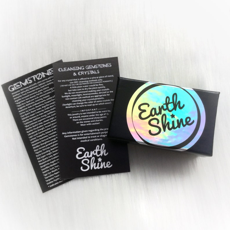 May include: A black box with a holographic label that reads "Earth Shine" is next to two black cards with white text. The cards list gemstone properties and cleansing instructions. The overall theme is related to gemstones and crystals.