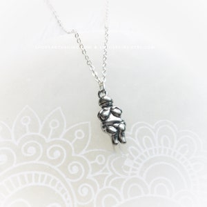 May include: Silver necklace with a charm of a small, round, silver figure with arms crossed over its chest. The figure is wearing a hat.