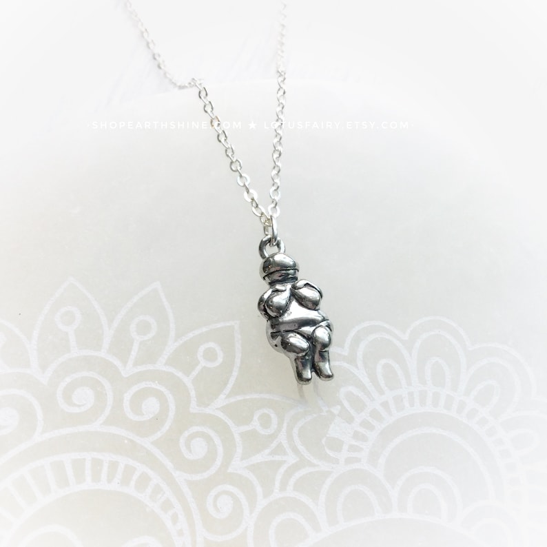 May include: Silver necklace with a charm of a small, round, silver figure with arms crossed over its chest. The figure is wearing a hat.