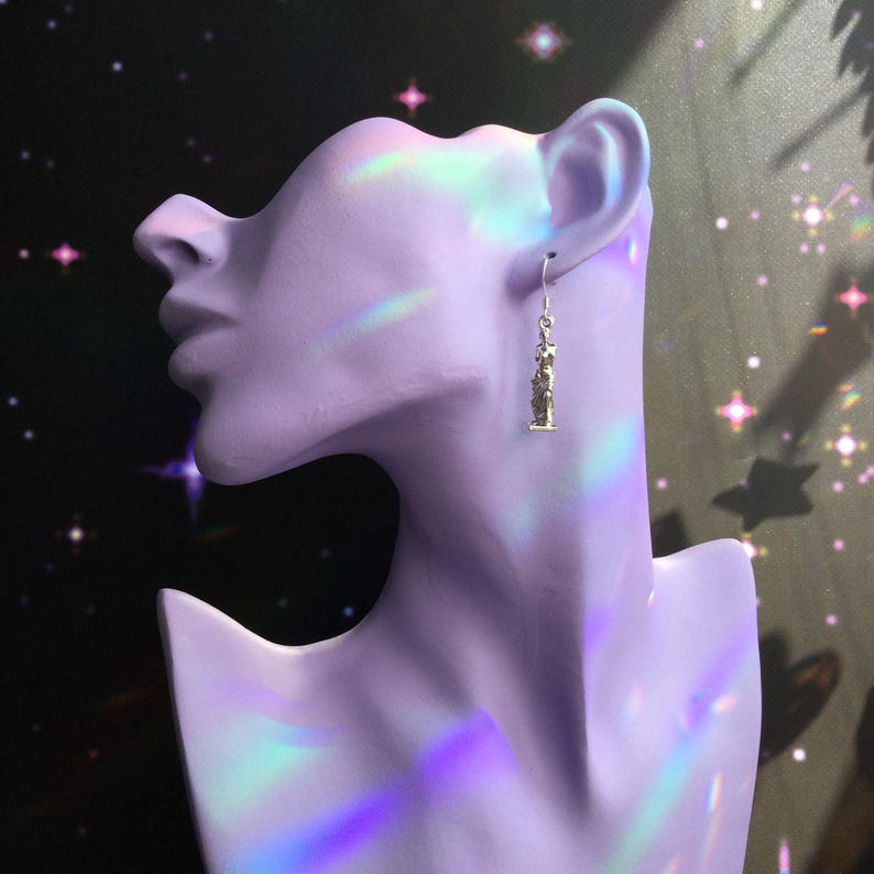 May include: A single gold-colored earring with a statue design hangs from a silver hook. The earring is displayed on a lavender bust against a dark background with star-shaped accents.