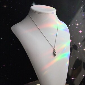 May include: A silver necklace with a small, detailed pendant shaped like a person. The necklace is displayed on a white mannequin bust against a black background with white stars.