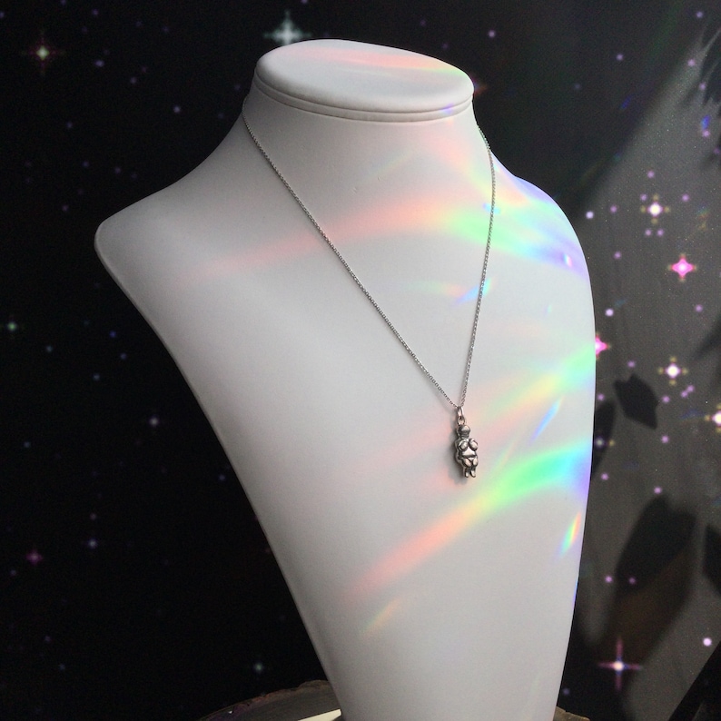 May include: A silver necklace with a small, detailed pendant shaped like a person. The necklace is displayed on a white mannequin bust against a black background with white stars.