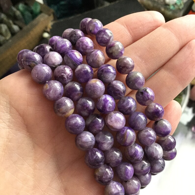 May include: A collection of purple gemstone bead bracelets. The beads are round and have a marbled appearance with shades of lavender, violet, and white. The bracelets are held in a hand, showcasing the natural variations in the stone.