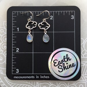 May include: Silver cloud-shaped earrings with teardrop-shaped, iridescent moonstone pendants. The earrings are displayed on a black surface with inch measurements. A circular sticker with the text "Earth Shine" is in the lower right corner.