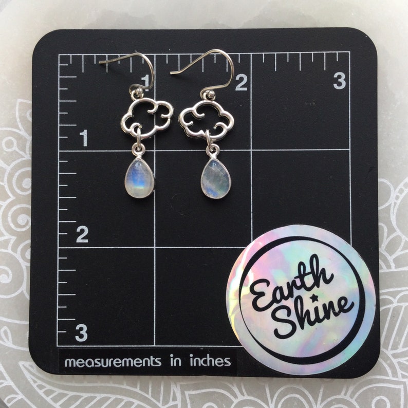 May include: Silver cloud-shaped earrings with teardrop-shaped, iridescent moonstone pendants. The earrings are displayed on a black surface with inch measurements. A circular sticker with the text "Earth Shine" is in the lower right corner.