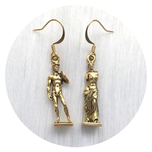 May include: Gold-toned dangle earrings featuring miniature sculptures of classical figures. One earring depicts a male statue, the other a female statue. The earrings have a hook closure and are set against a white background.