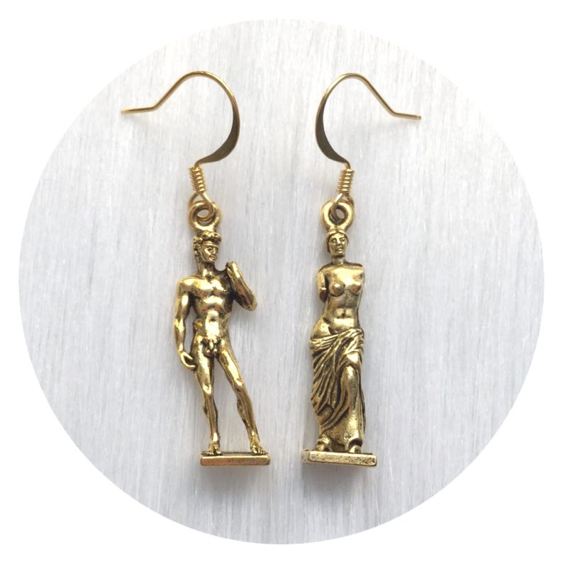 May include: Gold-toned dangle earrings featuring miniature sculptures of classical figures. One earring depicts a male statue, the other a female statue. The earrings have a hook closure and are set against a white background.