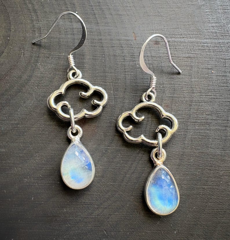 May include: A pair of silver earrings. Each earring has a cloud-shaped top and a teardrop-shaped, iridescent, blue and white stone pendant. The earrings have a hook closure.