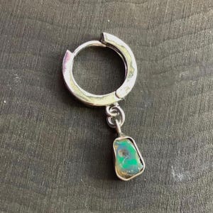 May include: A single silver hoop earring with a small, rectangular, iridescent green and blue stone pendant. The stone is set in a silver frame and hangs from a delicate chain. The earring is displayed on a dark, textured surface.