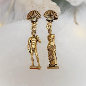 May include: Gold-toned dangle earrings featuring miniature sculptures. One earring depicts a male figure, the other a draped female figure. Each figure hangs from a seashell-shaped top. The earrings have a vintage aesthetic.