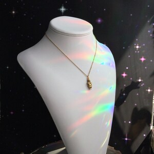 May include: A gold necklace with a small, detailed pendant shaped like a person. The necklace is displayed on a white mannequin against a black background with iridescent light reflecting off the necklace.