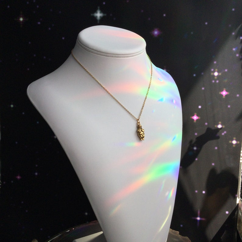 May include: A gold necklace with a small, detailed pendant shaped like a person. The necklace is displayed on a white mannequin against a black background with iridescent light reflecting off the necklace.