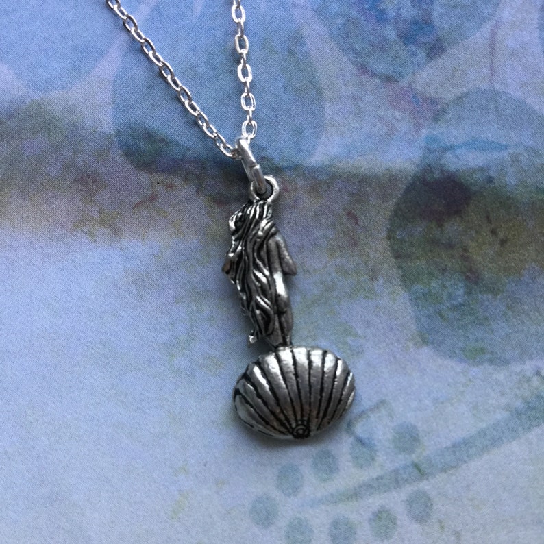 May include: A silver necklace with a pendant featuring a woman standing on a seashell. The pendant is detailed and has a vintage look.