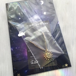 May include: A gold necklace with a sun and moon pendant. The pendant has a face on the sun and a crescent moon shape. The necklace is packaged in a clear plastic bag with a black background and white stars. The bag is labeled "Jewelry Care".